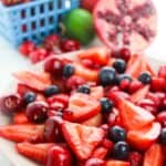 A vibrant features fresh sliced strawberries, cherries, blueberries, and raspberries, with a basket of strawberries and a halved pomegranate in the background.