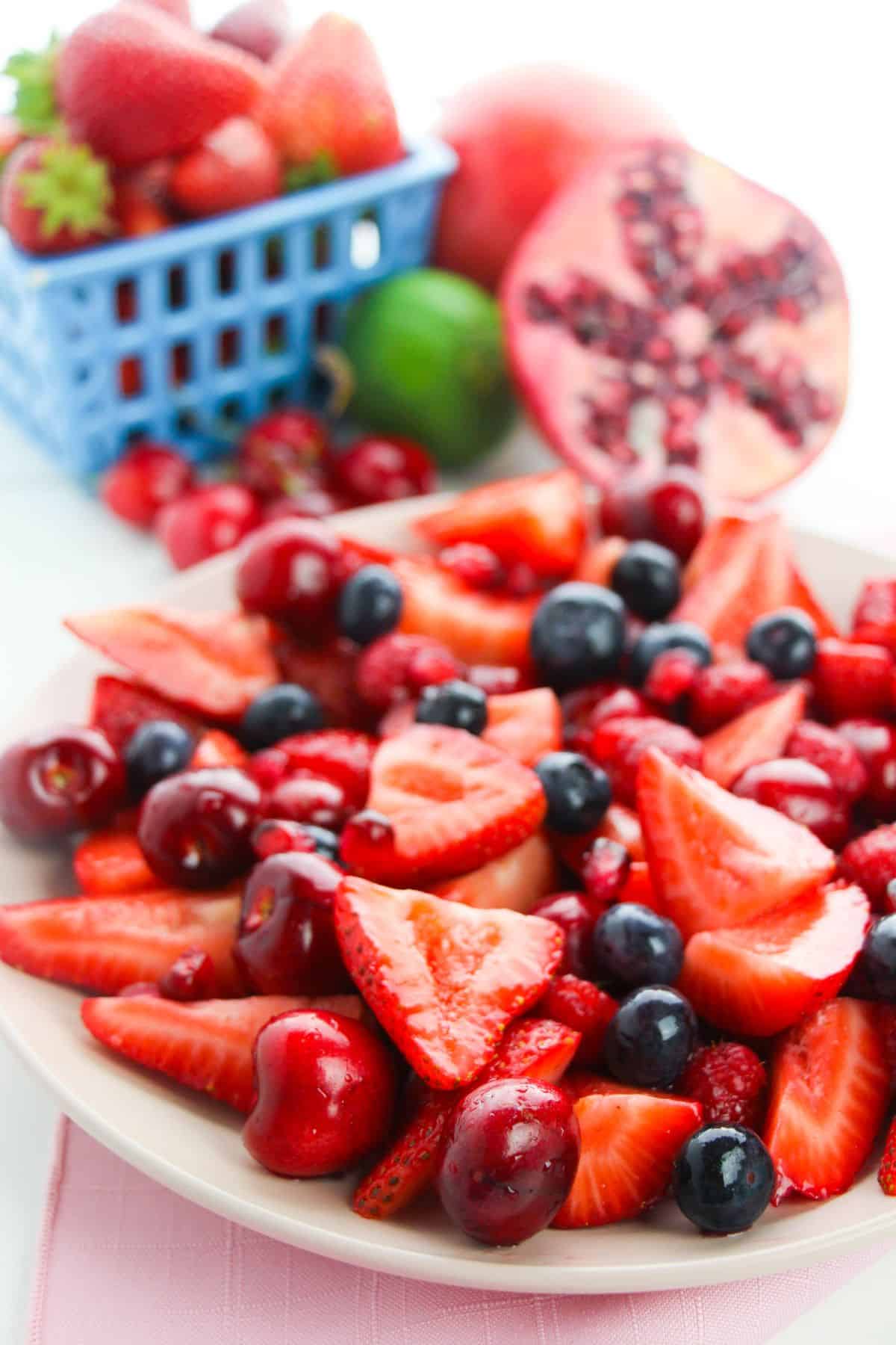A vibrant  features fresh sliced strawberries, cherries, blueberries, and raspberries, with a basket of strawberries and a halved pomegranate in the background.