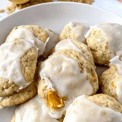 A plate of Peach Cookies with visible peach chunks is perfectly frosted. Brown sugar and sliced peaches are displayed in the background.