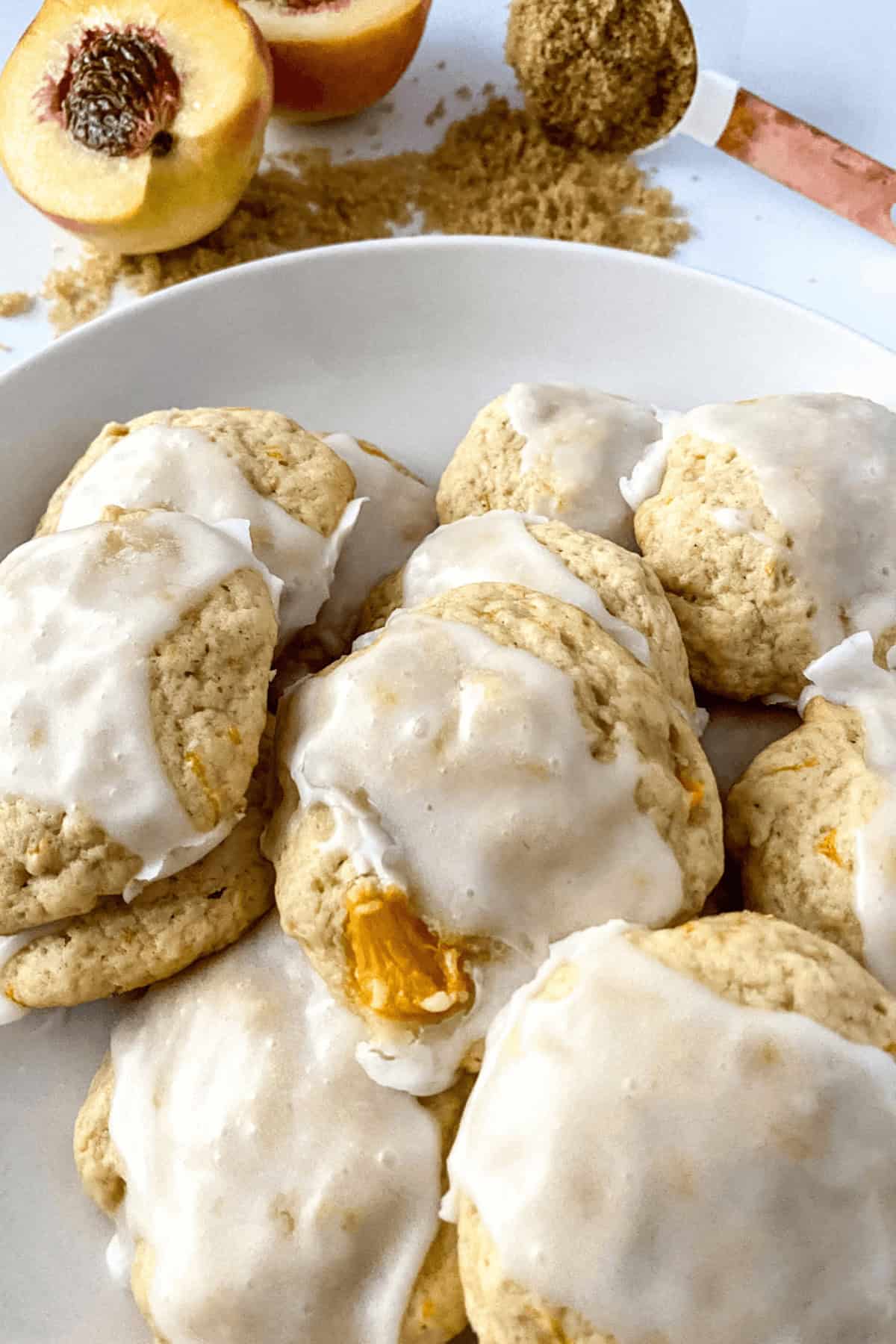 A plate of Peach Cookies with visible peach chunks is perfectly frosted. Brown sugar and sliced peaches are displayed in the background.