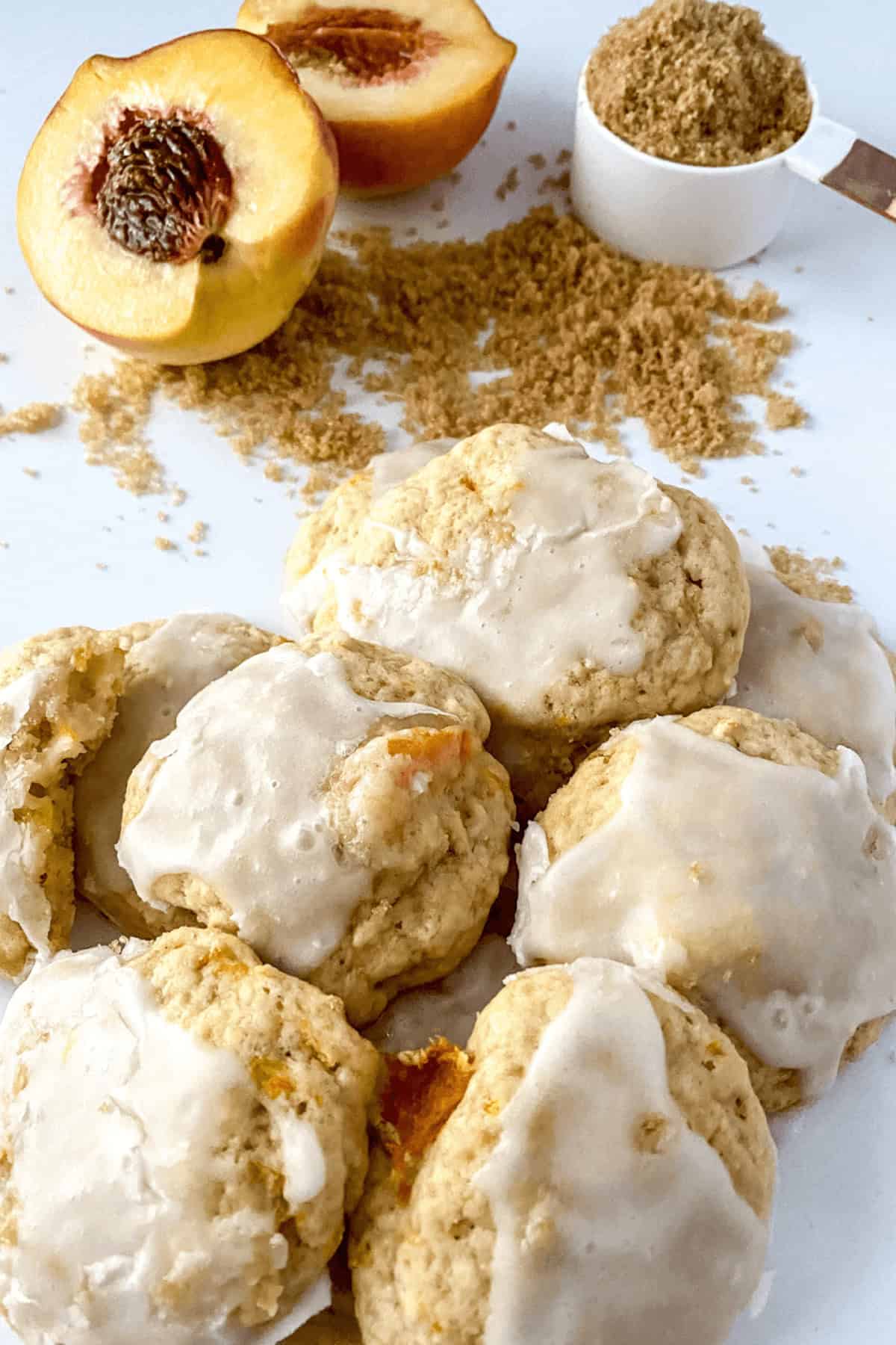 The image showcases a group of scrumptious peach cookies on a white surface. In the background, there is a halved peach and a measuring cup filled with brown sugar.