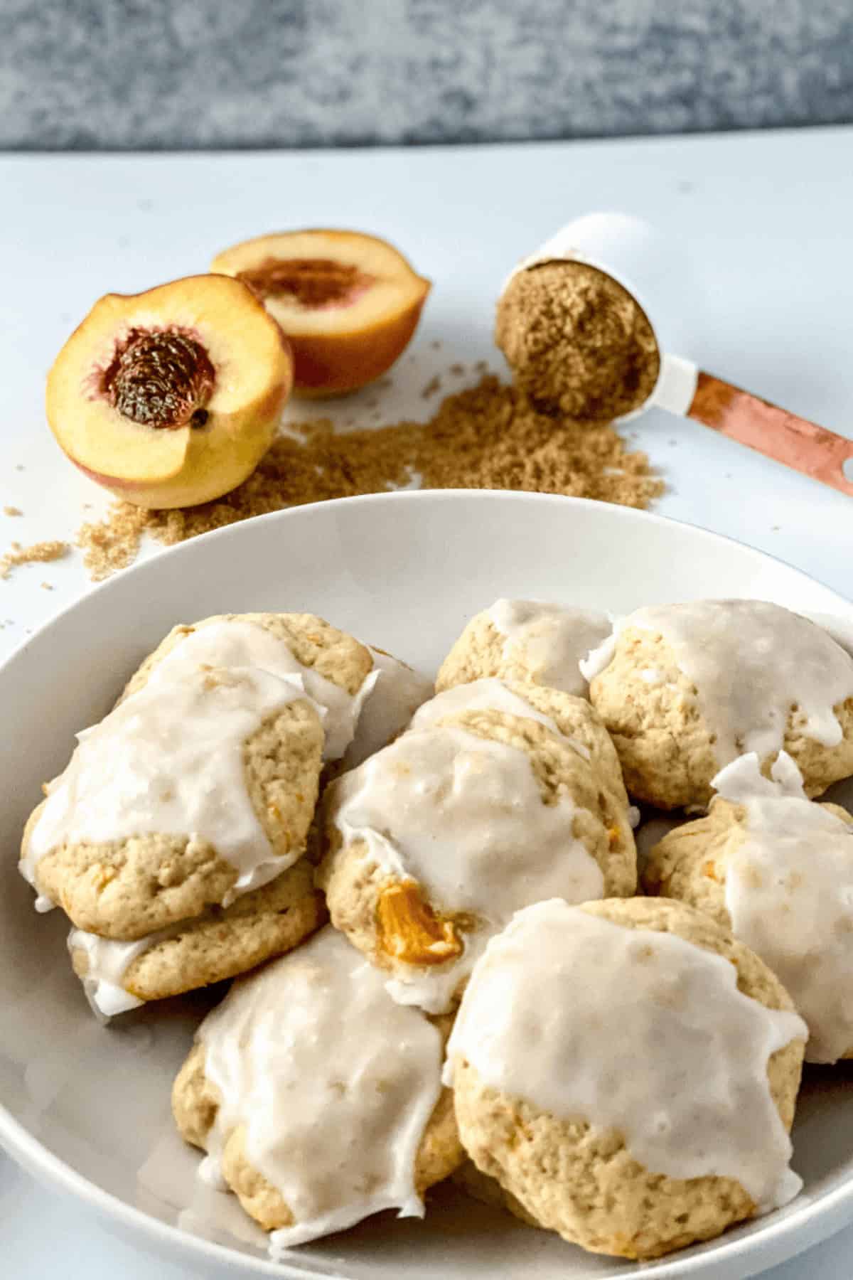 A bowl of frosted peach cookies is in the foreground, with a sliced peach and a measuring cup of brown sugar in the background.