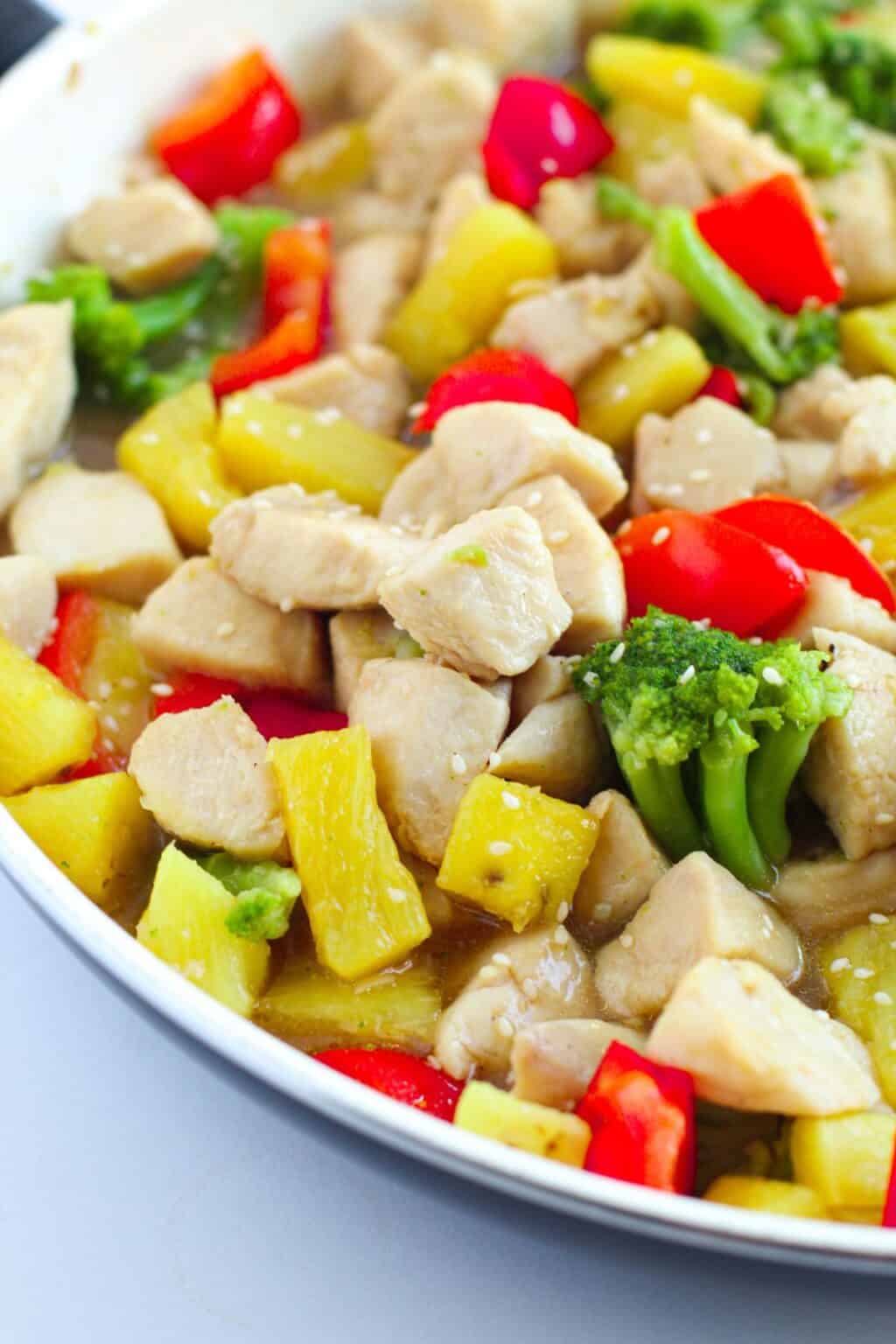 A close-up of Pineapple Chicken Stir Fry with pieces of chicken, red bell peppers, pineapple chunks, and broccoli in a white pan.