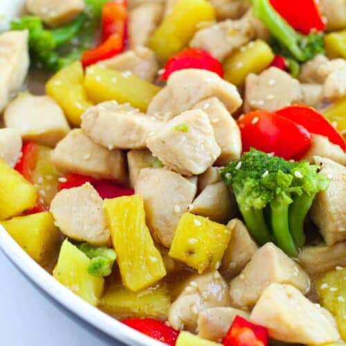 A close-up of Pineapple Chicken Stir Fry with pieces of chicken, red bell peppers, pineapple chunks, and broccoli in a white pan.
