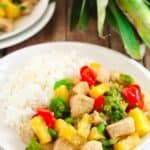 A white plate with fluffy rice and Pineapple Chicken Stir Fry—featuring chicken, broccoli, red bell pepper, and pineapple chunks—rests on a wooden table beside a fresh pineapple.