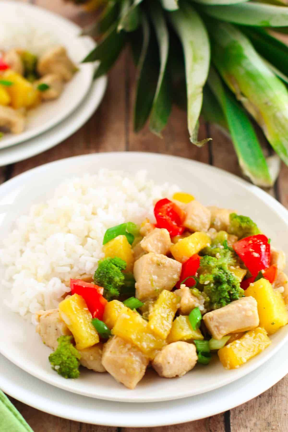A white plate with fluffy rice and Pineapple Chicken Stir Fry—featuring chicken, broccoli, red bell pepper, and pineapple chunks—rests on a wooden table beside a fresh pineapple.