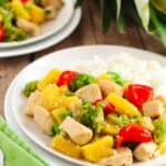 A plate of Pineapple Chicken Stir Fry with broccoli, red bell peppers, pineapple chunks, and white rice sits on a wooden table with a green napkin.