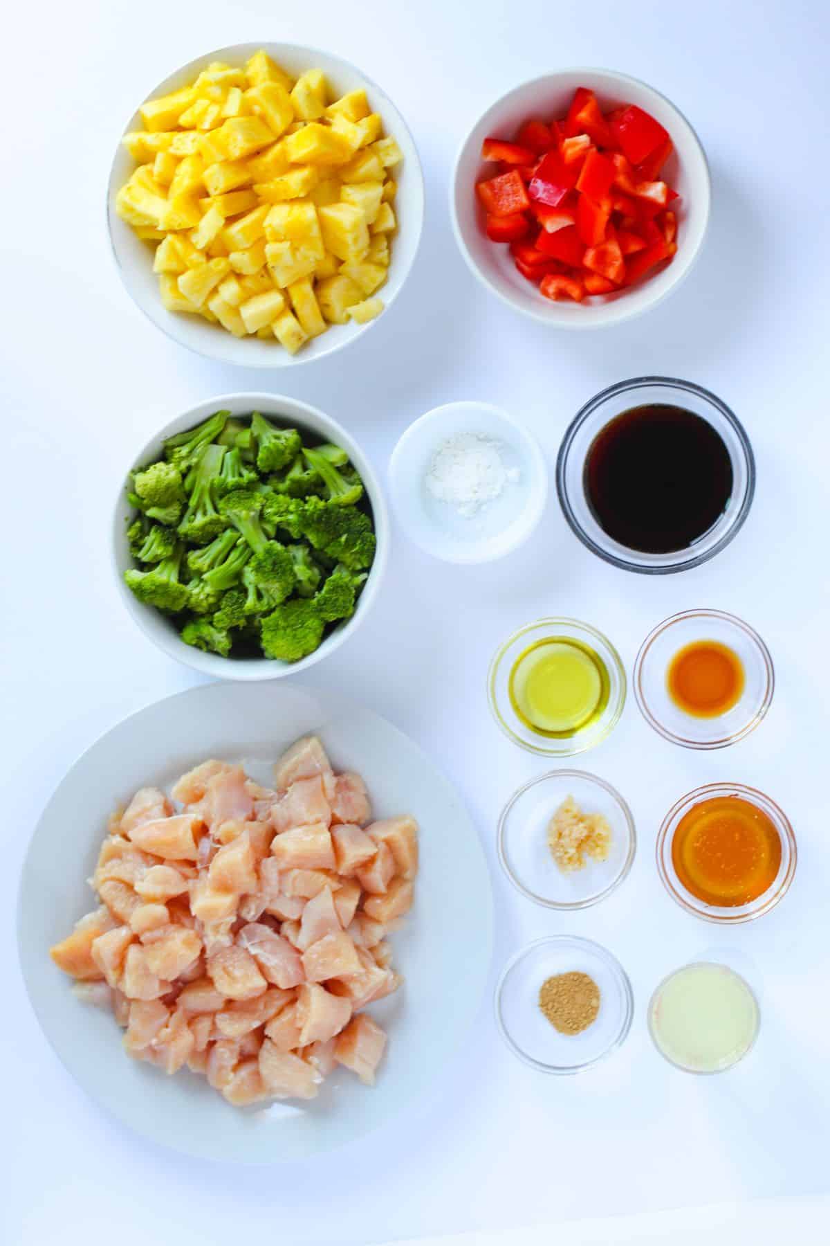 Overhead view of bowls with ingredients chopped pineapple, red bell pepper, broccoli, diced raw chicken, cornstarch, soy sauce, oil, honey, minced garlic, ginger, and lemon juice on a white background.