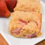 Close-up of two pieces of strawberry lemon crumb bars, now dubbed Strawberry Lemon Blondies, on a white plate with fresh strawberries and a lemon slice in the background.