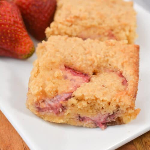 Close-up of two pieces of strawberry lemon crumb bars, now dubbed Strawberry Lemon Blondies, on a white plate with fresh strawberries and a lemon slice in the background.