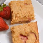 Two pieces of strawberry cake are served on a white plate with two whole strawberries on the side, reminiscent of the delightful flavors found in Strawberry Lemon Blondies.