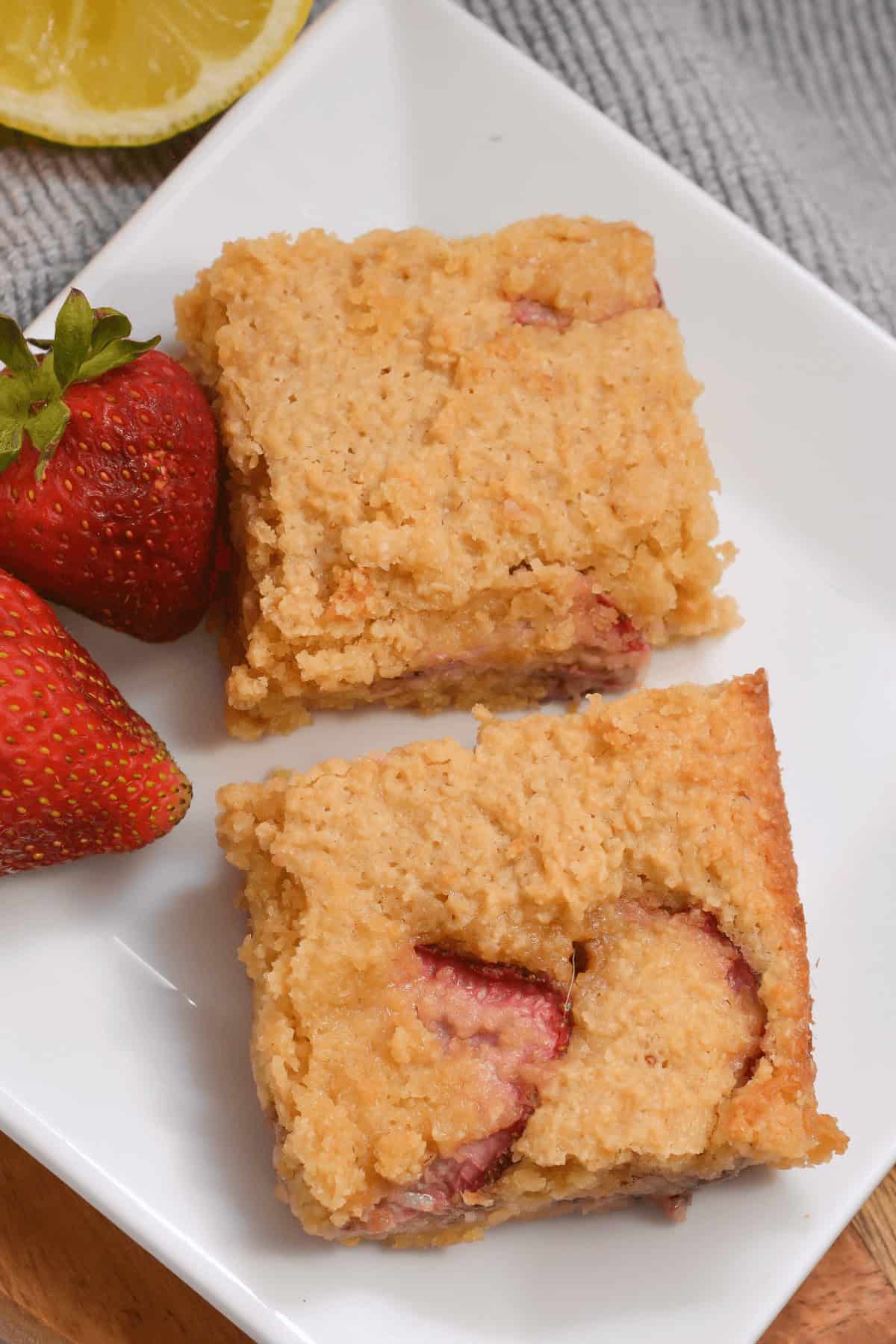 Two pieces of strawberry cake are served on a white plate with two whole strawberries on the side, reminiscent of the delightful flavors found in Strawberry Lemon Blondies.
