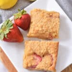 A white plate with two square slices of Strawberry Lemon Blondies, garnished with two whole strawberries. A half lemon and whole lemon are in the background on a gray cloth.