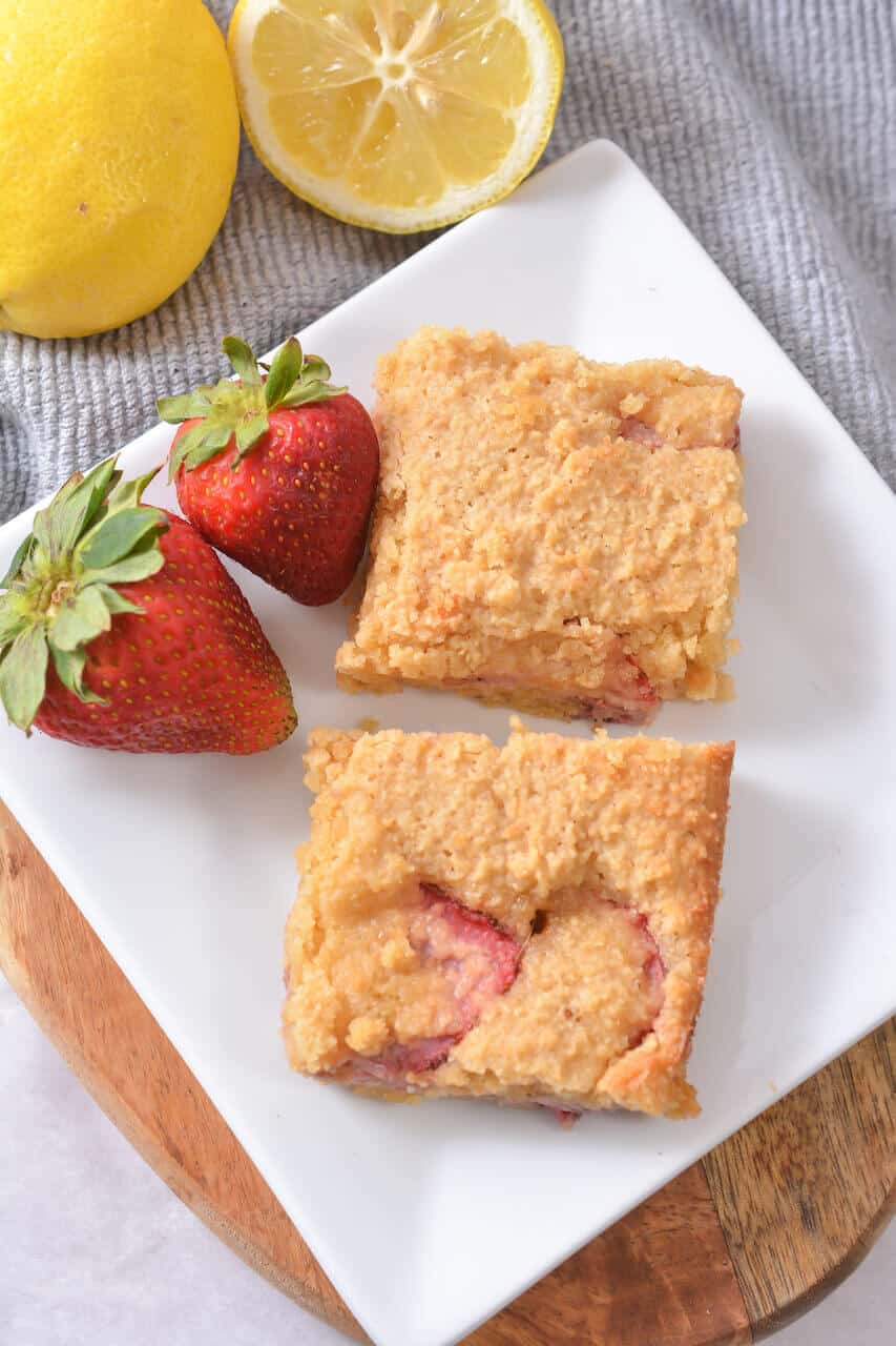A white plate with two square slices of Strawberry Lemon Blondies, garnished with two whole strawberries. A half lemon and whole lemon are in the background on a gray cloth.