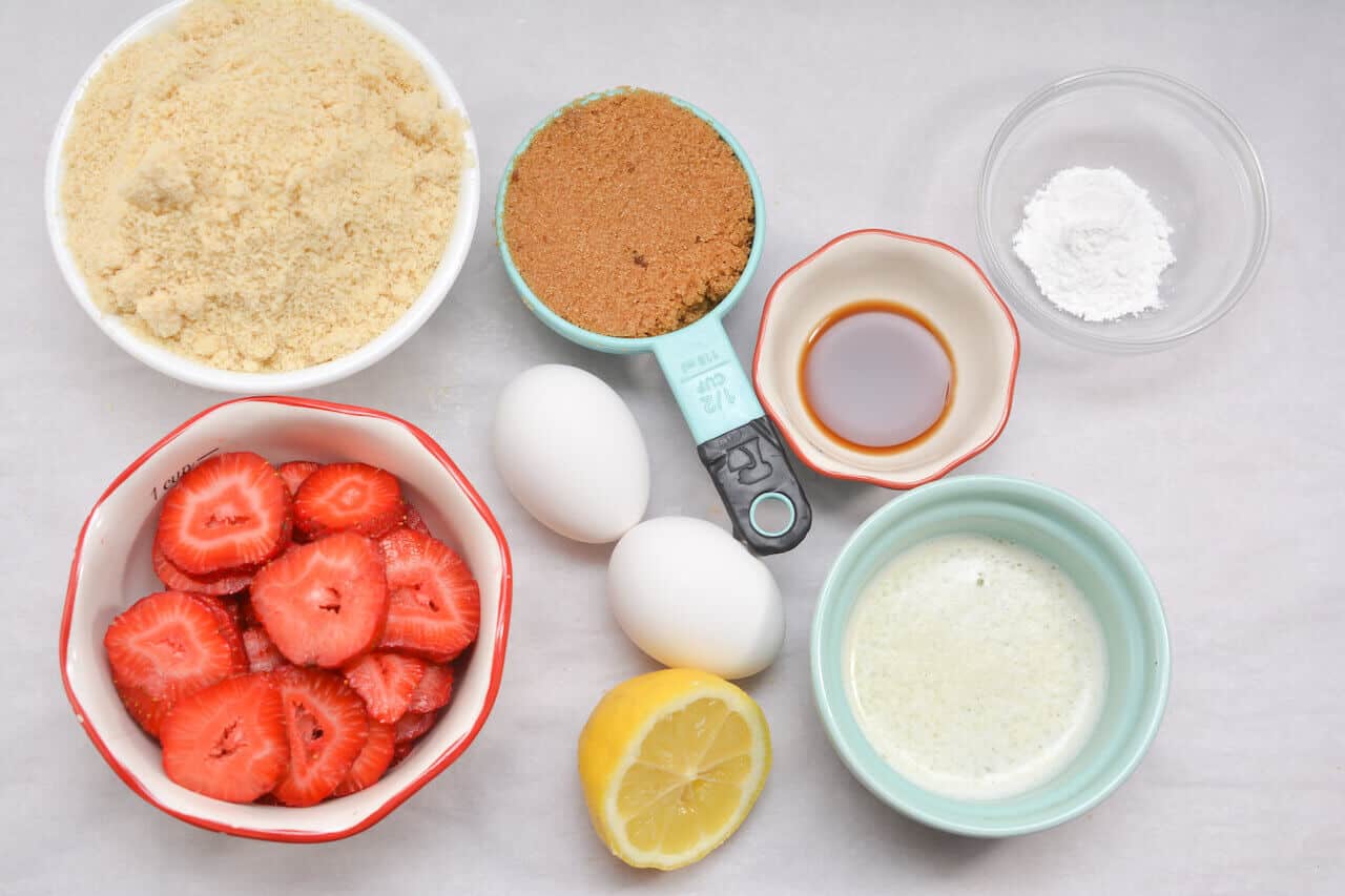 Ingredients for baking laid out on a surface: almond flour, brown sugar, baking powder, strawberries, two eggs, vanilla extract, melted butter, and a lemon. 