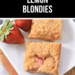Two Strawberry Lemon Blondies with visible strawberry pieces are served on a white plate, accompanied by whole strawberries and a lemon in the background.