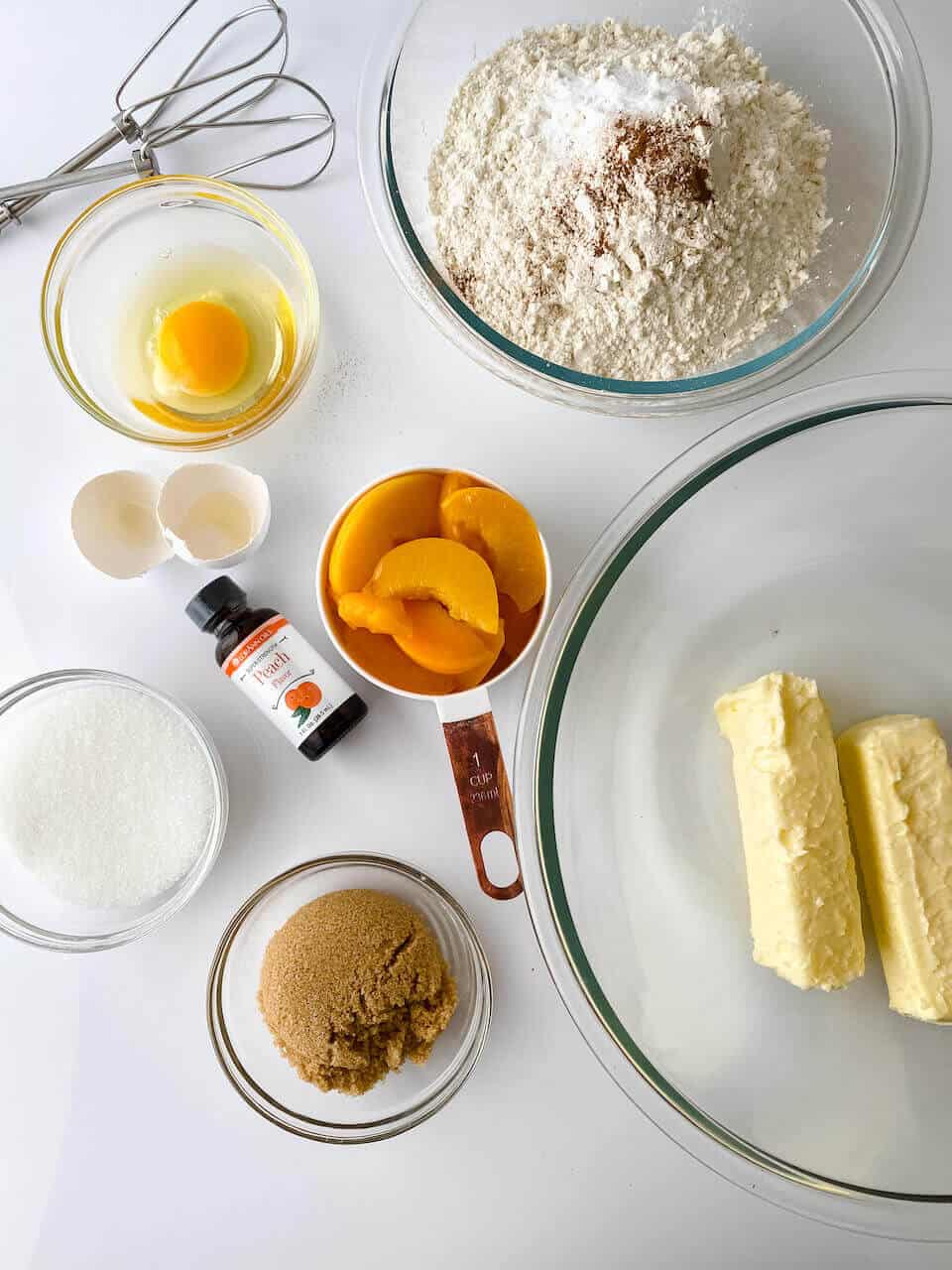 Ingredients for baking Peach Cookies including flour, sugar, brown sugar, butter, eggs, sliced peaches, vanilla extract, and mixing beaters arranged on a white surface.