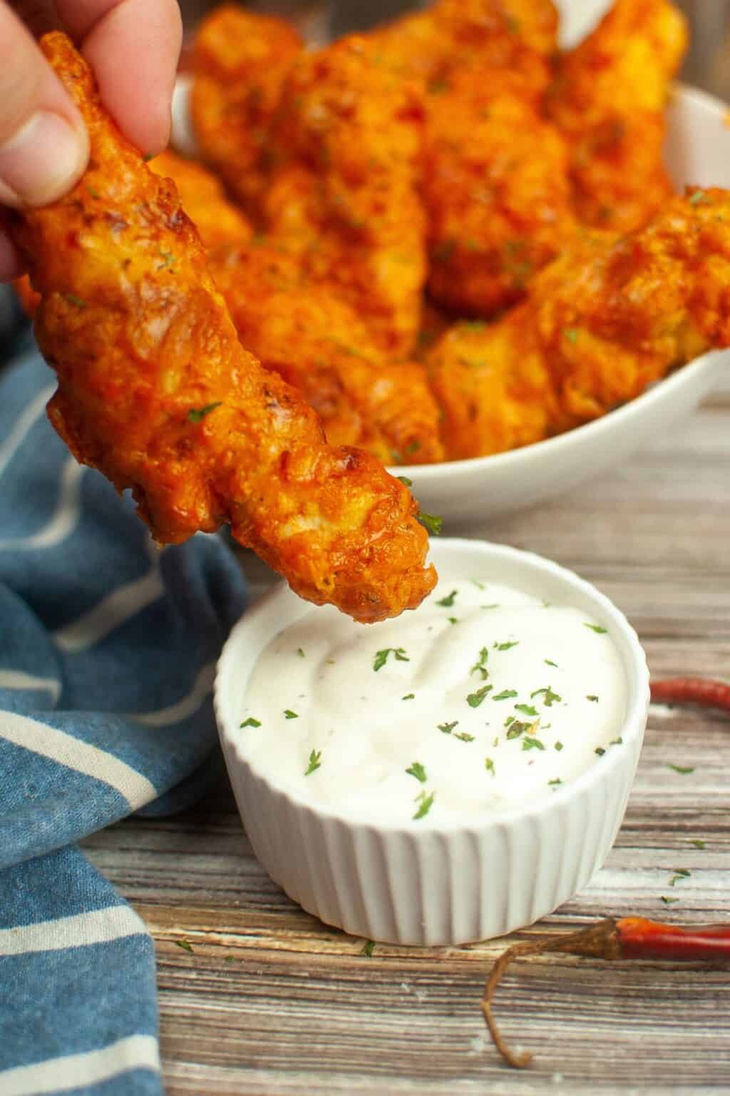 A hand dips a piece of crispy Air Fryer Buffalo Chicken into a small bowl of white sauce, with more chicken pieces in a bowl in the background.