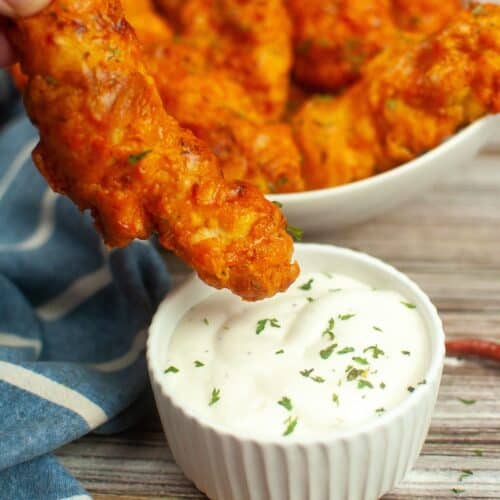 A hand dips a piece of crispy Air Fryer Buffalo Chicken into a small bowl of white sauce, with more chicken pieces in a bowl in the background.