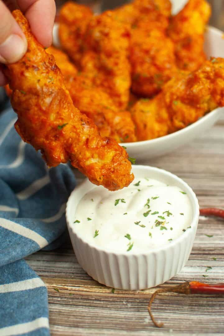 A hand dips a piece of crispy Air Fryer Buffalo Chicken into a small bowl of white sauce, with more chicken pieces in a bowl in the background.