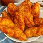 A white bowl filled with several pieces of crispy Air Fryer Buffalo Chicken wings coated in a reddish-orange sauce, placed on a wooden surface next to a blue striped cloth.