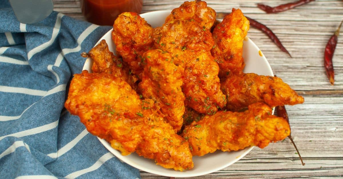 A white bowl filled with several pieces of crispy Air Fryer Buffalo Chicken wings coated in a reddish-orange sauce, placed on a wooden surface next to a blue striped cloth.