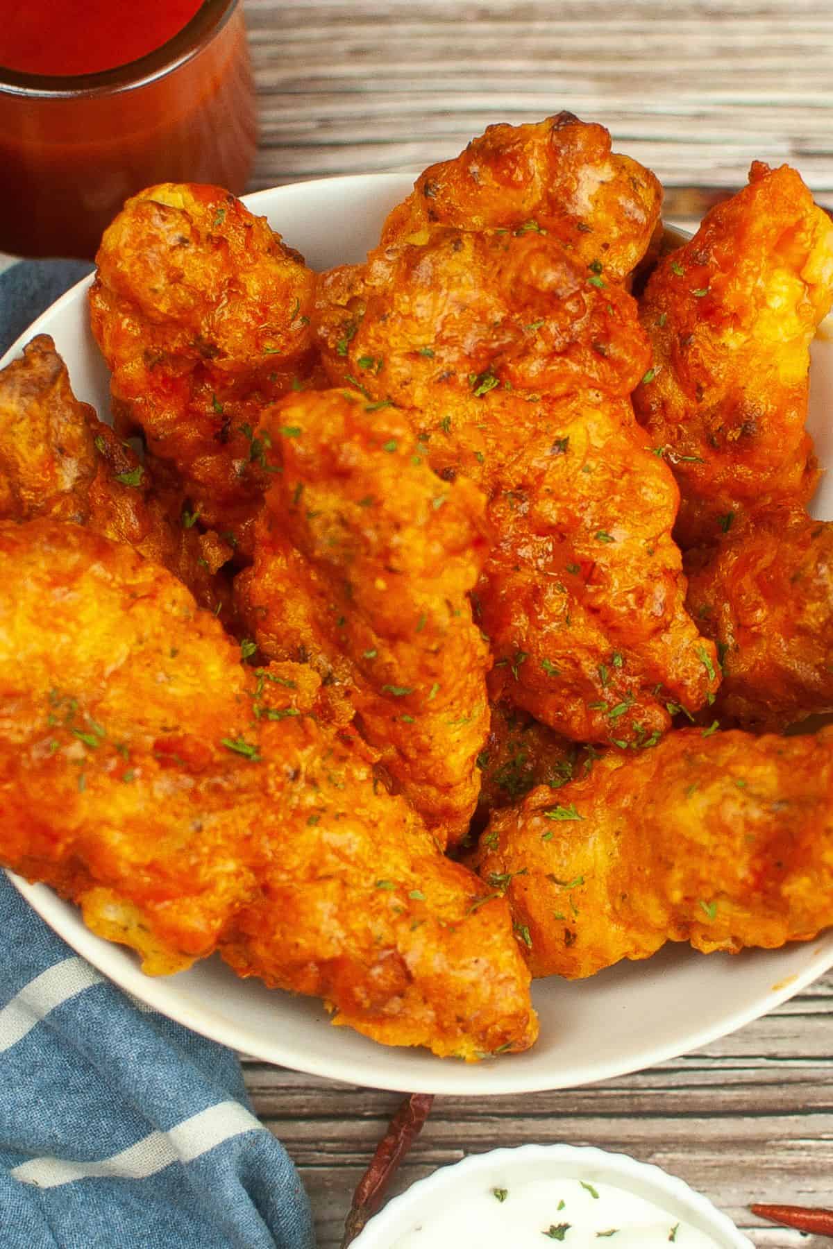 A bowl of crispy strips is placed on a wooden surface with a blue napkin and a cup of dipping sauce nearby.