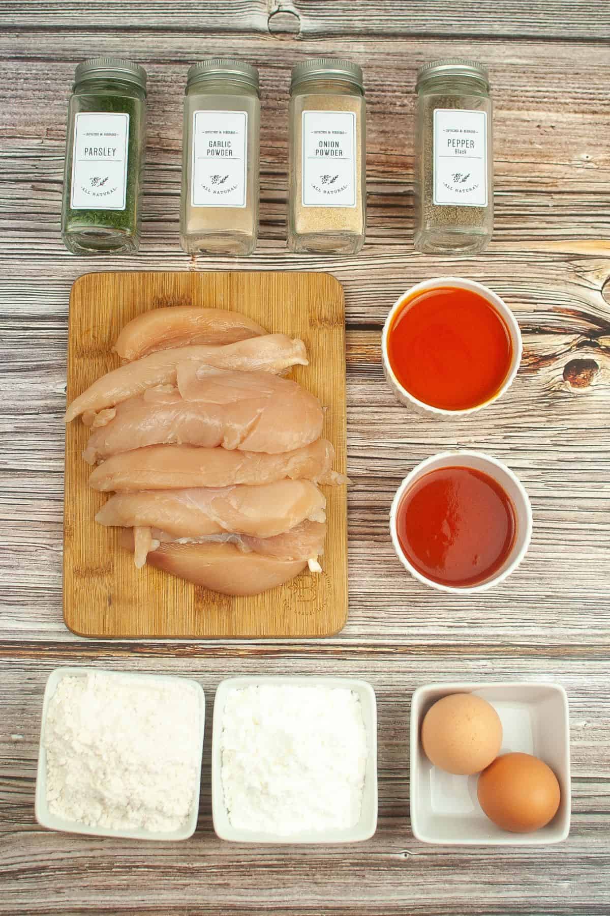 Raw chicken strips on a wooden board, surrounded by bowls of flour, cornstarch, eggs, hot sauce—perfect for Air Fryer Buffalo Chicken—and jars of parsley, onion powder, garlic powder, and pepper on a wooden surface.