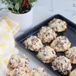 A black baking tray with Almond Joy Cookies—filled with coconut and chocolate chips—rests on a marble counter, beside a yellow patterned cloth and a small potted plant with green leaves and red berries.