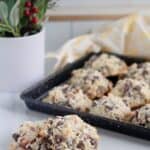 A tray of freshly baked Almond Joy Cookies with chocolate chips and nuts sits on a marble counter; a potted plant is in the background.