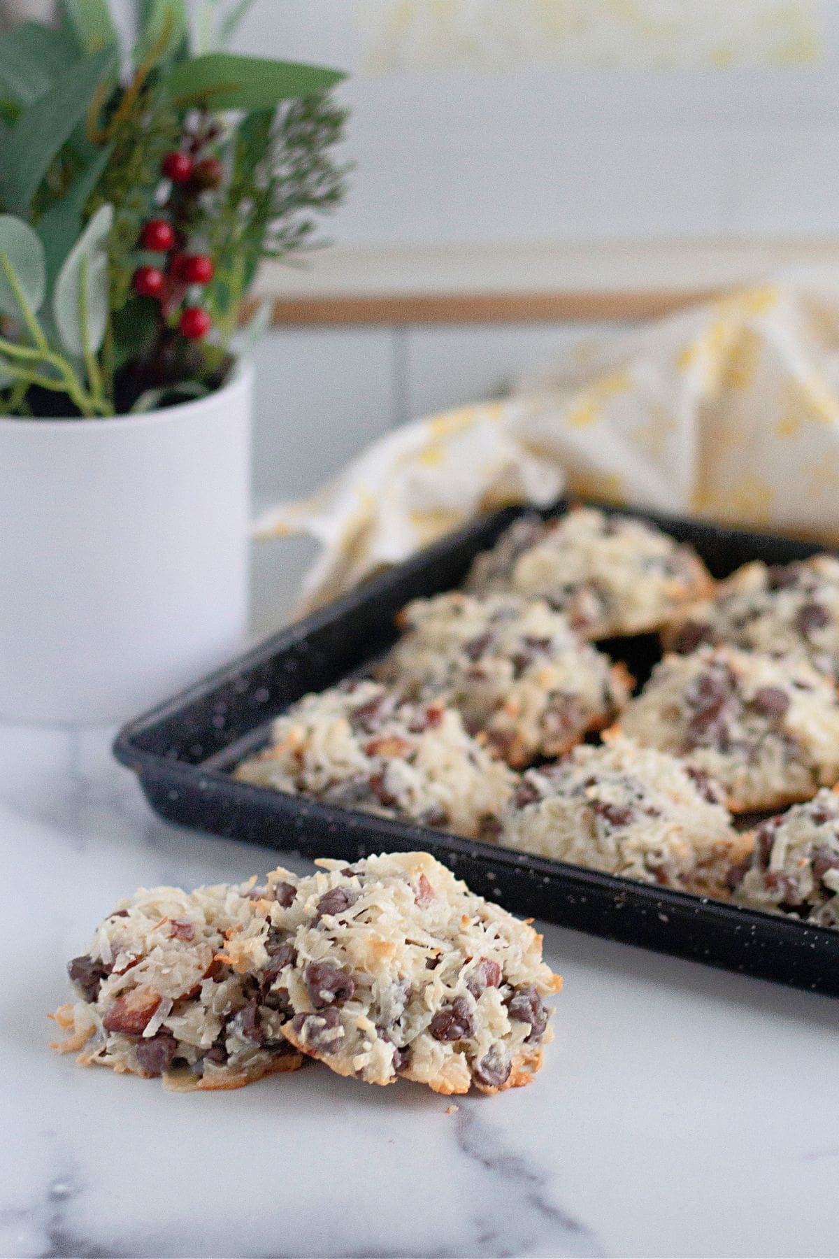 A tray of freshly baked Almond Joy Cookies with chocolate chips and nuts sits on a marble counter; a potted plant is in the background.