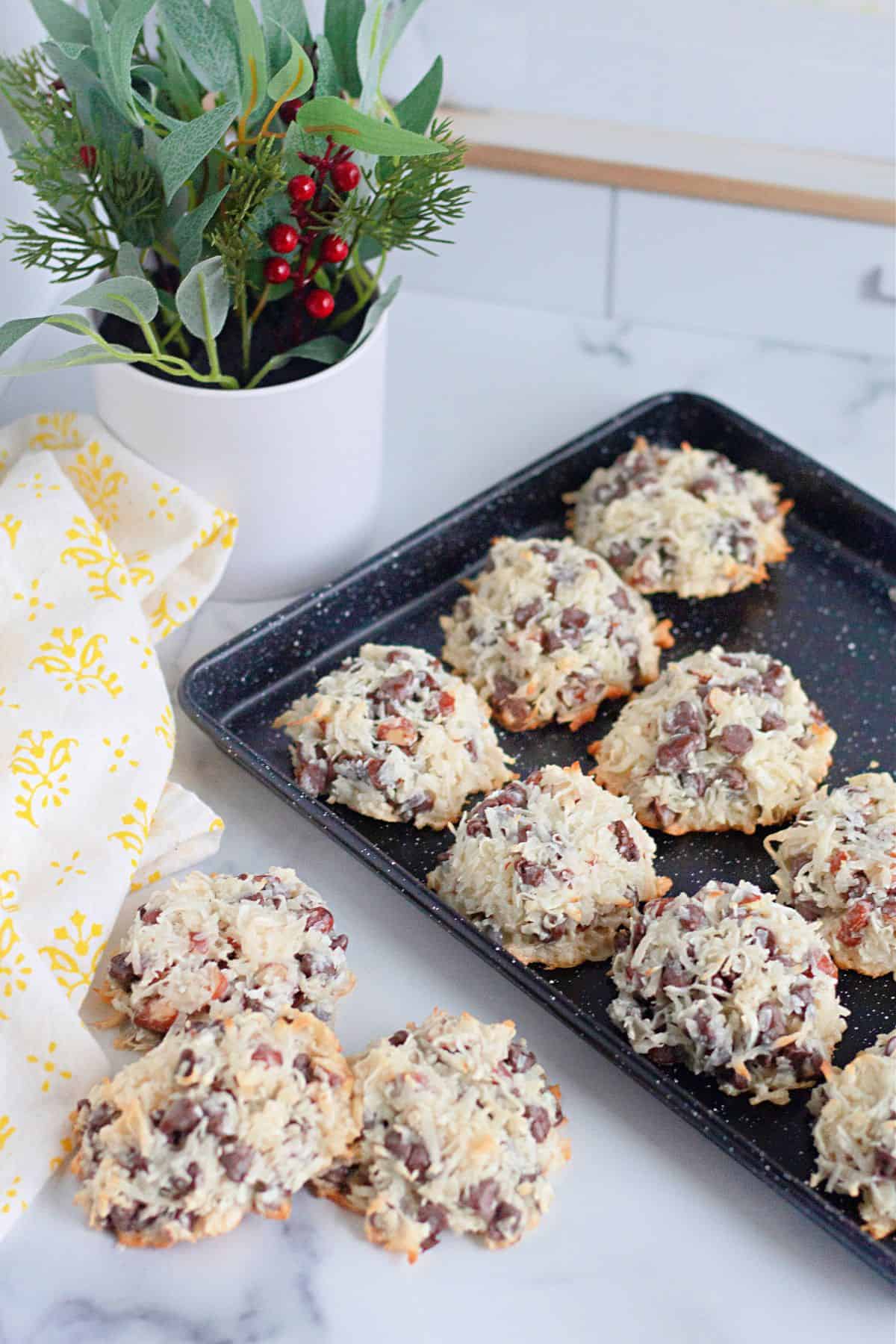 A black baking tray featuring coconut and chocolate chips, sits on a marble surface alongside a small potted plant and a yellow-patterned cloth.