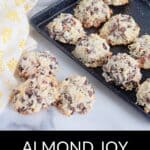 A tray and countertop display several Almond Joy Cookies made with coconut, chocolate chips, and nuts. A yellow-patterned cloth and a potted plant are also visible.