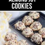 A black baking sheet with freshly baked Almond Joy Cookies—packed with chocolate chips, coconut, and almonds—rests on a marble surface beside a yellow patterned cloth.