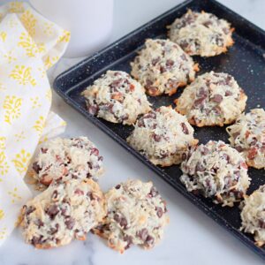 A black baking tray with Almond Joy Cookies—coconut and chocolate chip treats—rests nearby on a marble surface next to a yellow-patterned cloth.