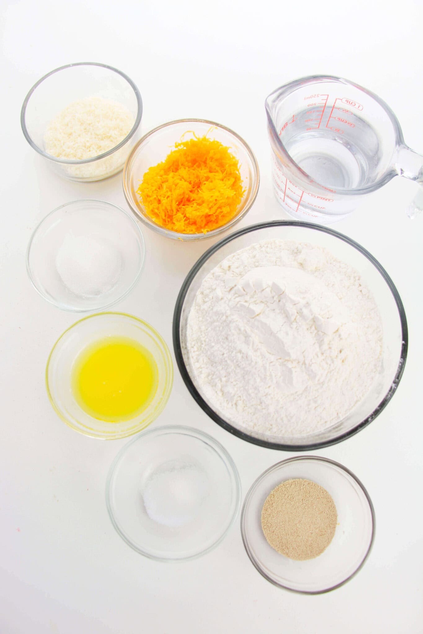 Various baking ingredients, perfect for making the snack including flour, orange zest, melted butter, water, yeast, sugar, and salt are measured in clear bowls on a white surface.