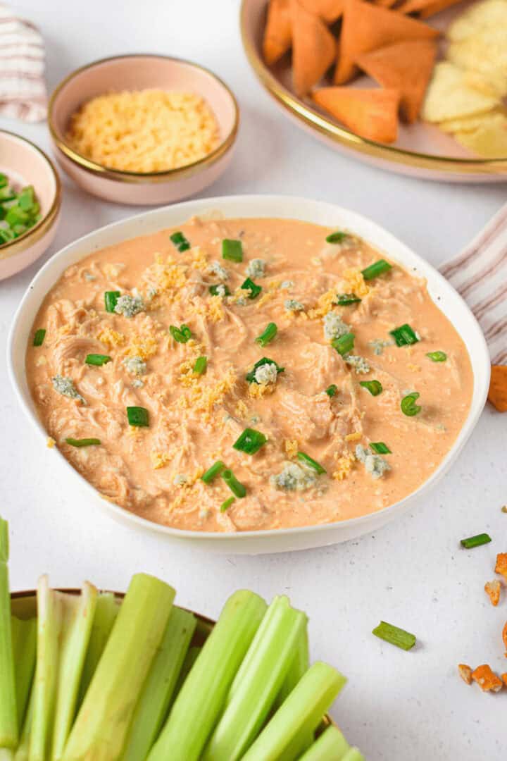 A bowl of creamy Instant Pot Buffalo chicken dip topped with chopped green onions and crumbled cheese. Celery sticks and triangular chips are visible nearby.