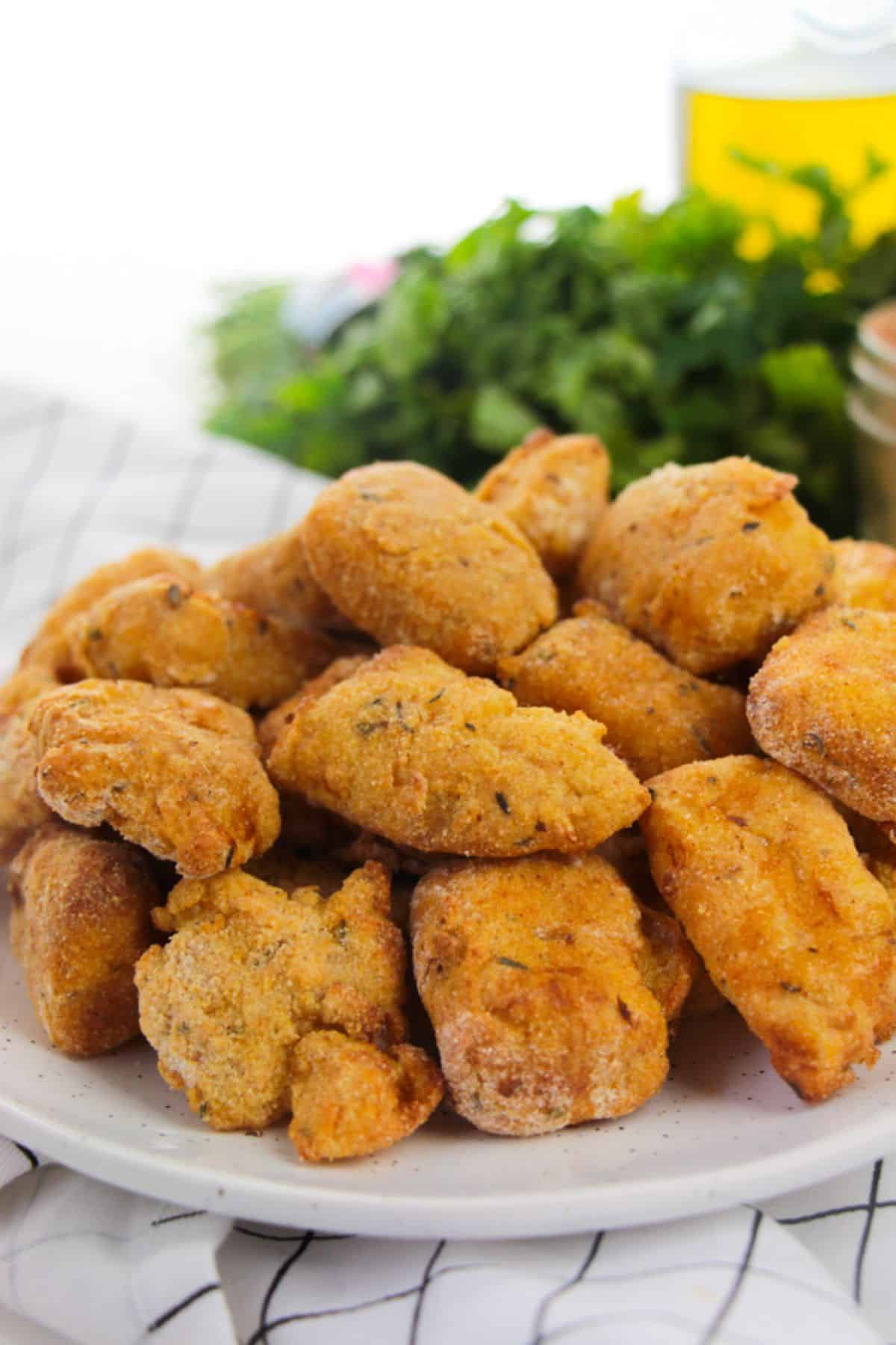 Plate of golden-brown, breaded Air Fryer Catfish Nuggets sits on a white plate, with a blurred background of fresh herbs and a checkered cloth.