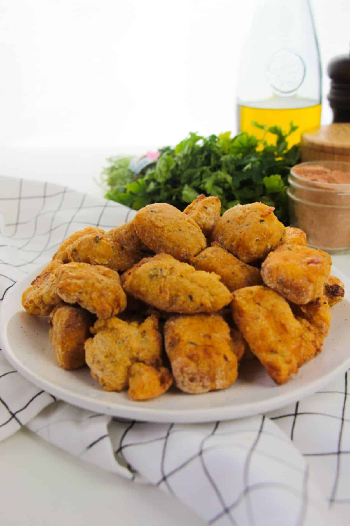 A plate of golden-brown, crispy Air Fryer Catfish Nuggets sits on a white napkin, with fresh herbs, seasoning, and a bottle in the background.