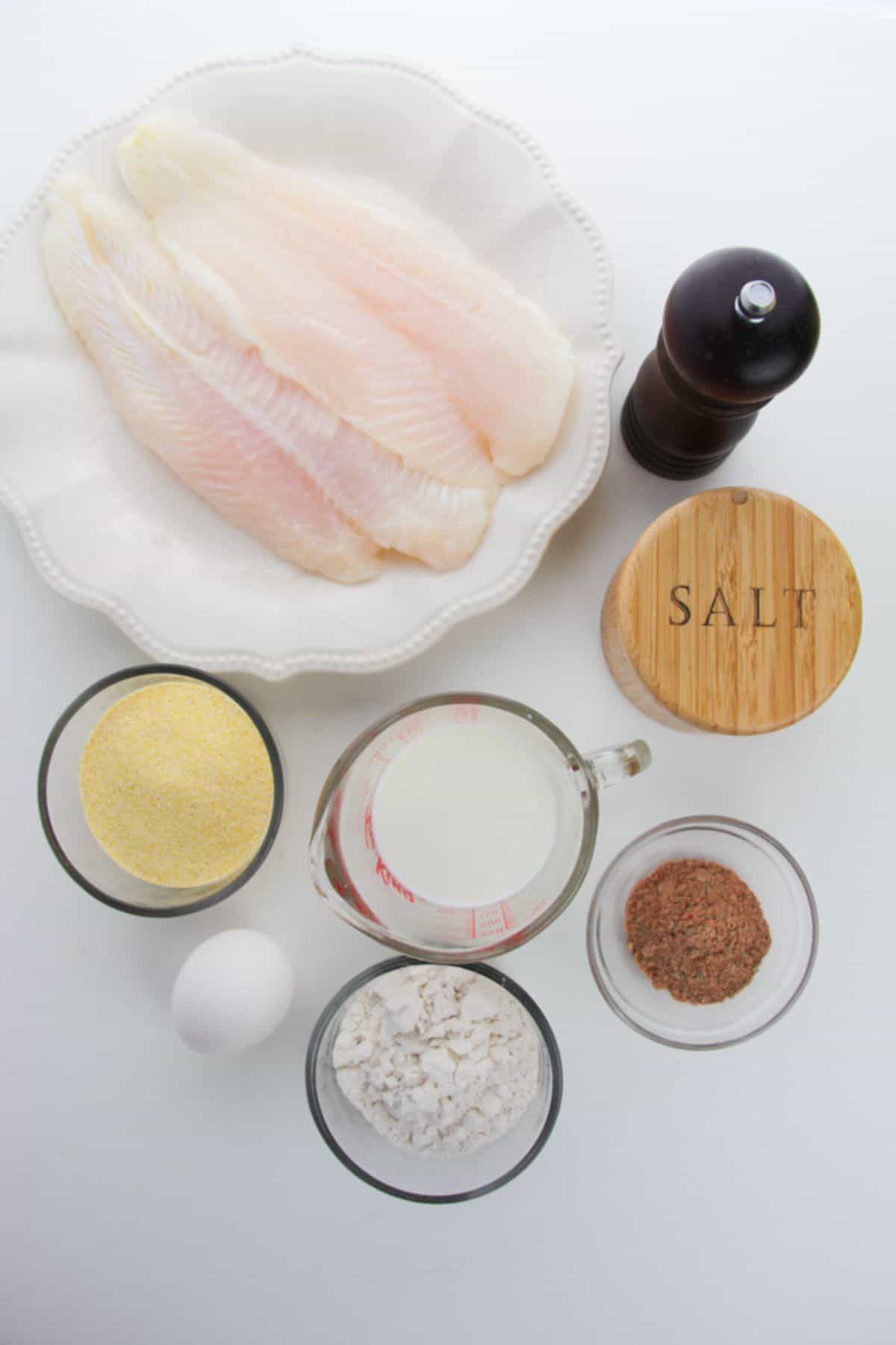 Raw fish fillets on a plate, next to a pepper grinder, salt container, cornmeal, milk, egg, flour, and ground spices in small bowls on a white background—everything you need to prepare delicious Air Fryer Catfish Nuggets.