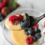 A spoon holding a raspberry and a blueberry covered in creamy yellow sauce, embodying the essence of berries and cream, with more berries and sauce visible in a blurred background.
