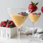 Two martini glasses filled with yellow custard topped with succulent berries, including strawberries, blueberries, and raspberries, on a white marble surface. A container of berries is in the background, adding to the Berries and Cream allure.