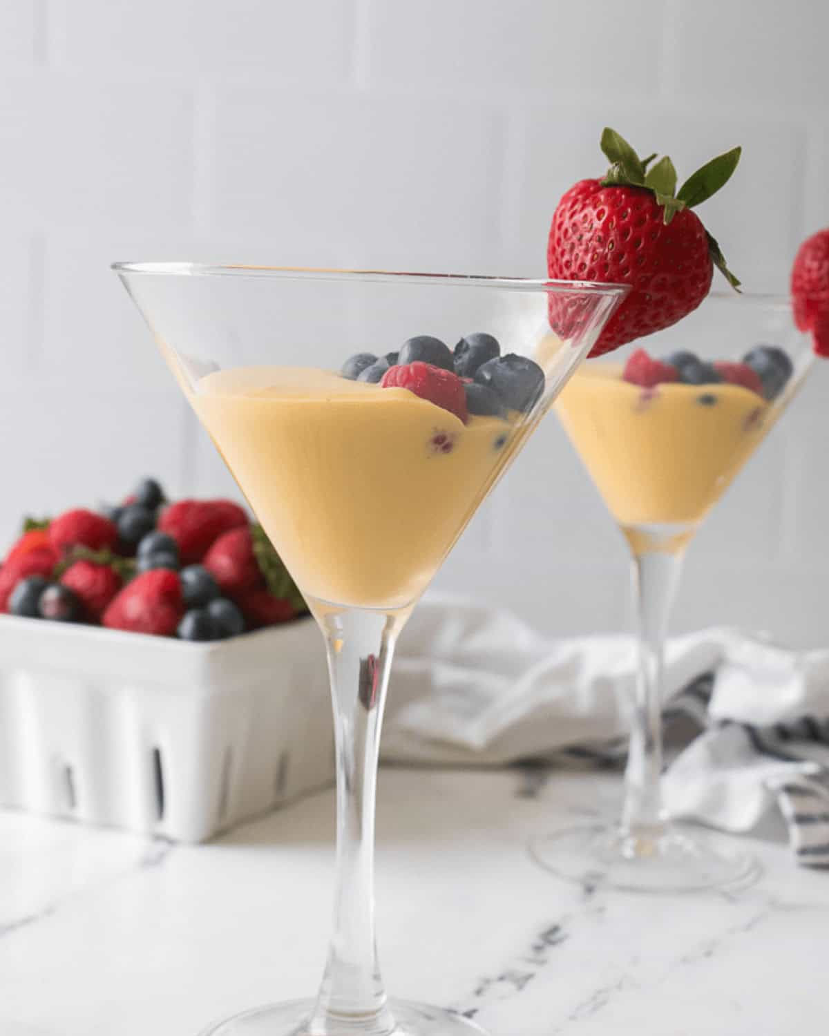 Two martini glasses filled with yellow custard topped with succulent berries, including strawberries, blueberries, and raspberries, on a white marble surface. A container of berries is in the background, adding to the Berries and Cream allure.