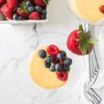 A glass bowl filled with yellow custard topped with raspberries and blueberries. A fresh strawberry sits on the rim. A bowl of mixed berries and a striped cloth napkin are in the background, evoking a perfect berries and cream delight.
