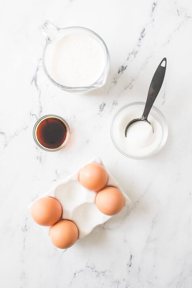 Items for baking on a marble surface: a glass measuring cup with milk, a small bowl with sugar and a measuring spoon, a container with vanilla extract, berries, and cream, and a carton with four brown eggs.