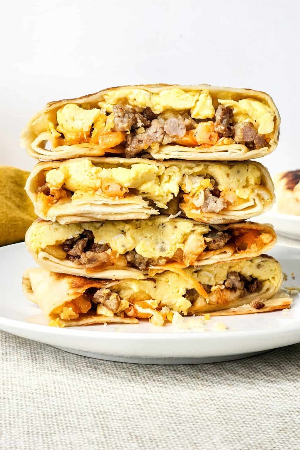 Three breakfast burritos filled with scrambled eggs, sausage, and cheese are stacked on a white plate against a plain background—perfect for anyone craving a hearty Breakfast Crunchwrap experience at home.