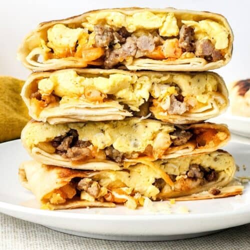 Three breakfast burritos filled with scrambled eggs, sausage, and cheese are stacked on a white plate against a plain background—perfect for anyone craving a hearty Breakfast Crunchwrap experience at home.