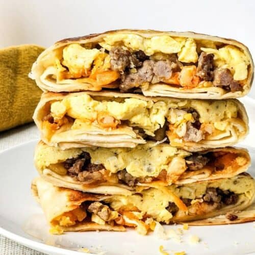 Three cut breakfast burritos filled with scrambled eggs, sausage, and cheese are stacked on a white plate, reminiscent of a classic Breakfast Crunchwrap; a glass of orange juice and a yellow napkin are in the background.
