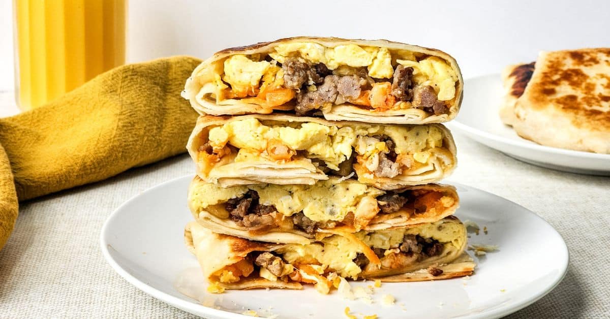 Stacked Breakfast Crunchwraps filled with scrambled eggs, sausage, and cheese on a white plate, with a glass of orange juice and a yellow cloth in the background.