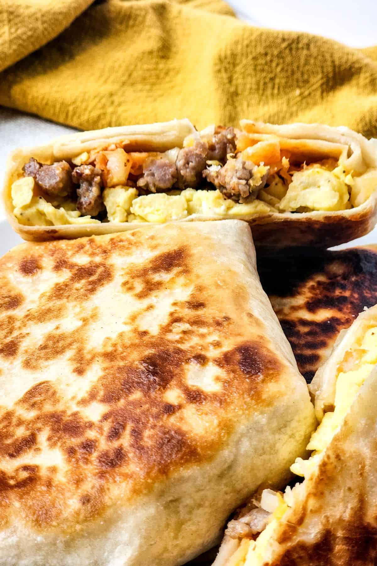 Close-up of folded and grilled Breakfast Crunchwrap burritos with scrambled eggs, sausage, cheese, and shredded vegetables, one cut in half to show the filling. A yellow cloth is in the background.